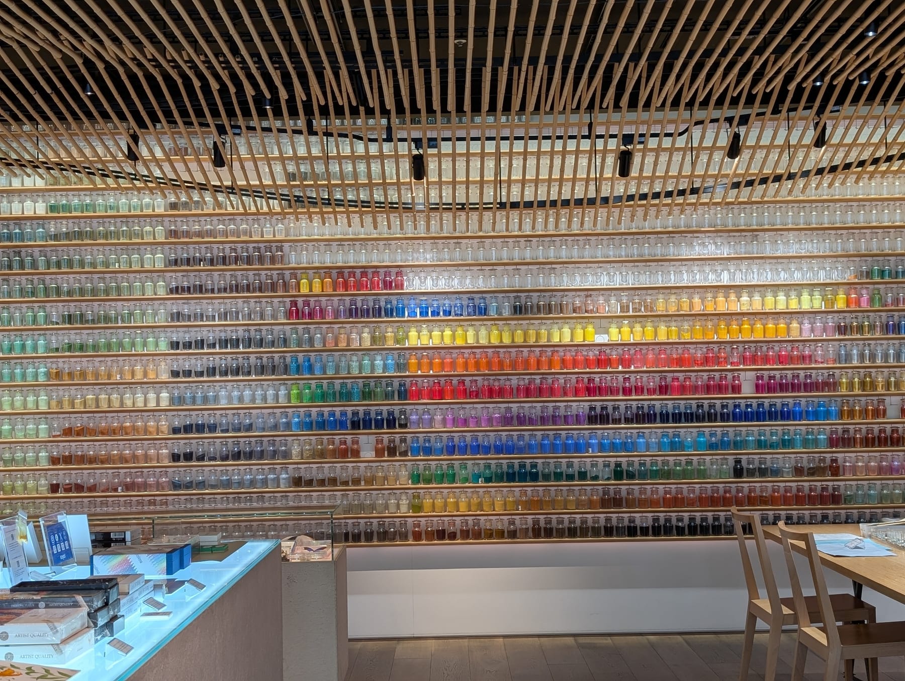 A shop wall of hundreds of small glass jars of powdered pigment, set in long rows and sorted by color from greens through yellows, oranges, reds, purples and blues, under a ceiling of narrow wooden slats.