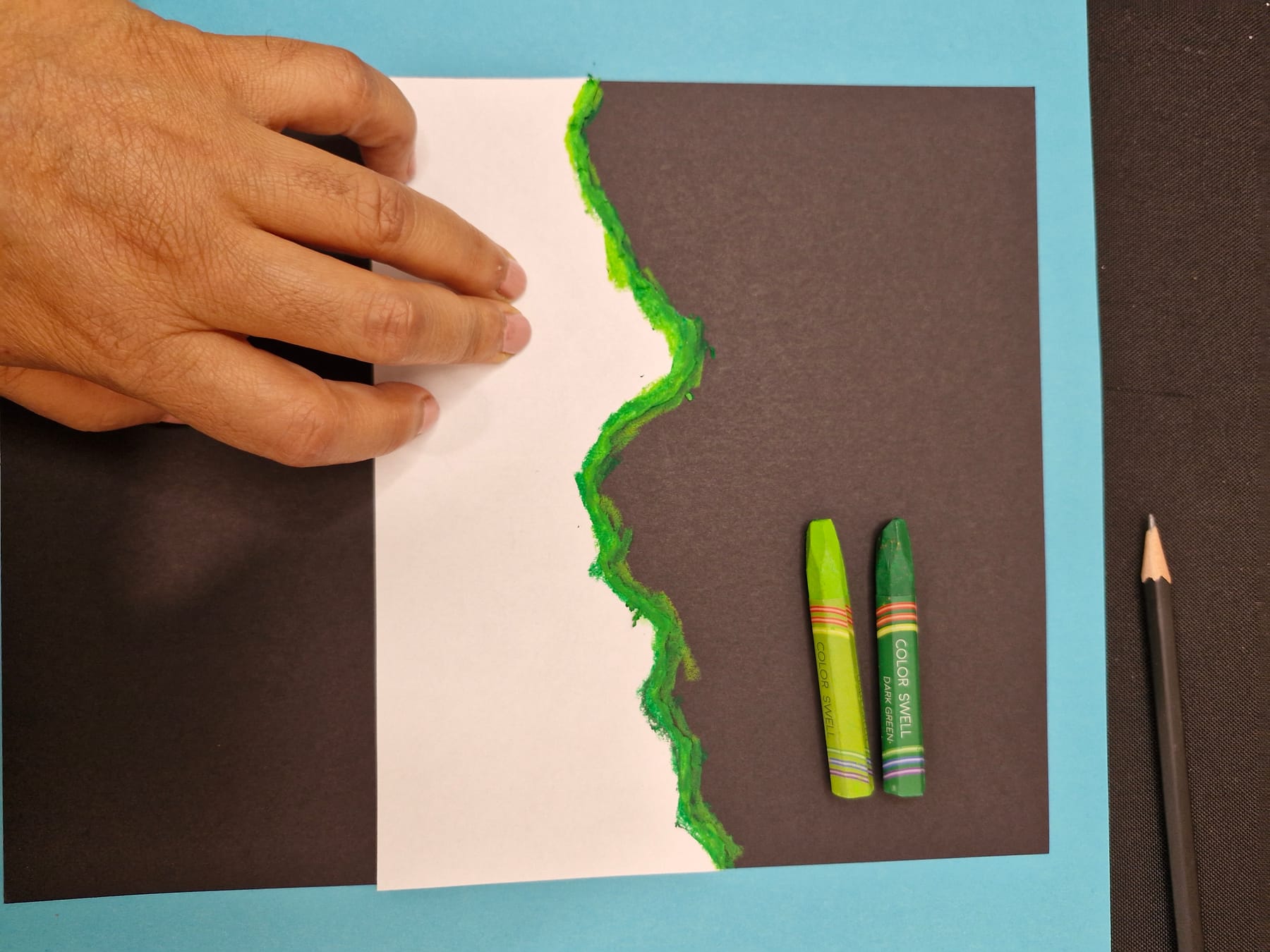 A torn edge colored with a thick band of green pastel, laid face down on the black paper and held flat by one hand.