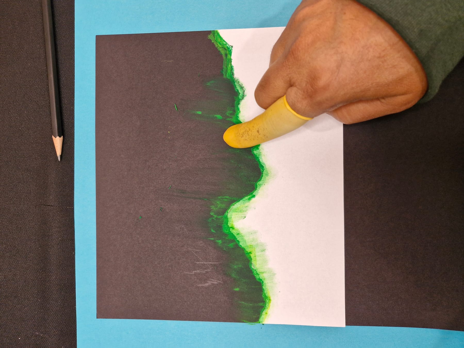 A fingertip rubbing the green pastel off the torn edge and out onto the black paper, away from the paper rather than along it.
