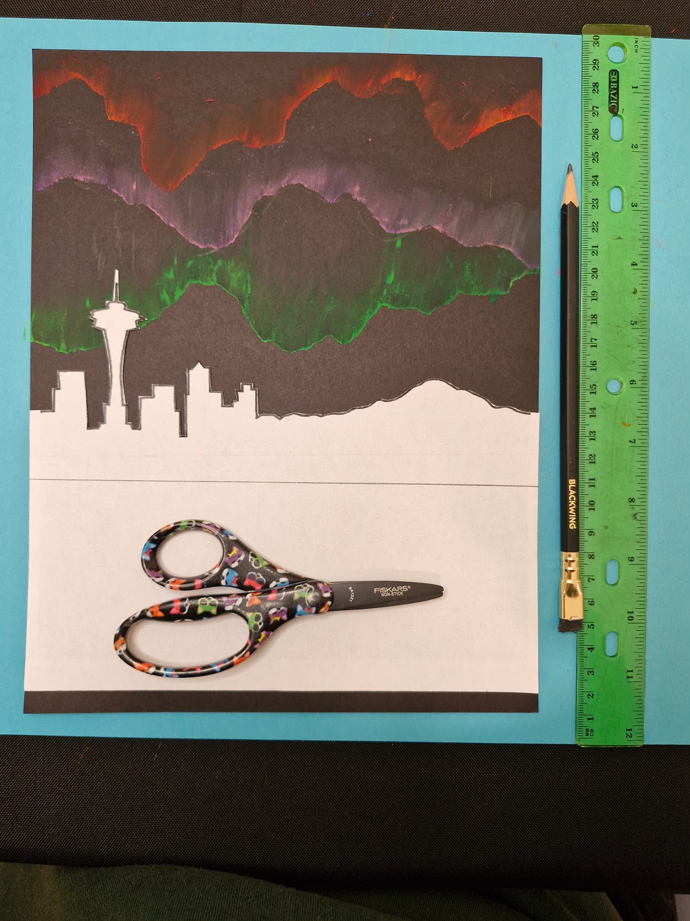 The cut skyline held against the finished aurora sky to try its position, with scissors and a ruler beside it.