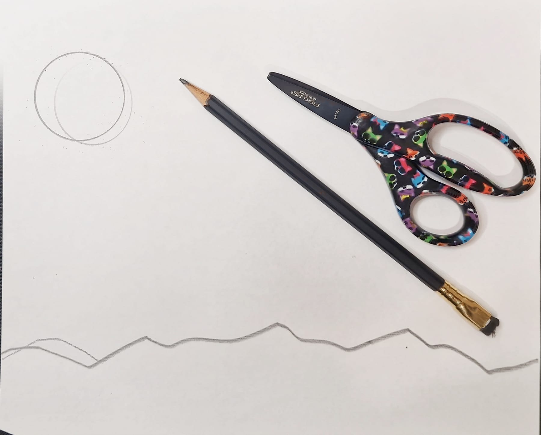 A sheet of white paper with a circle drawn in the top corner and a jagged mountain line across the bottom, with a pencil and scissors beside it.